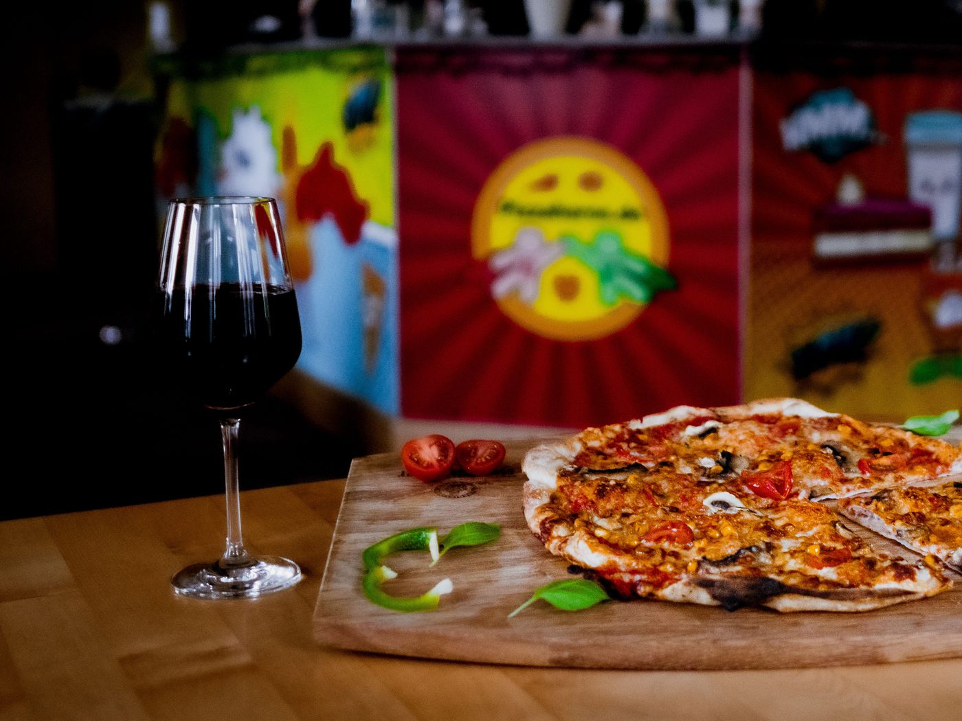 Gedeckter Tisch im Gastraum mit einer Pizza auf dem Holzbrett und einem Glas Rotwein vor der bunt bemalten Wand