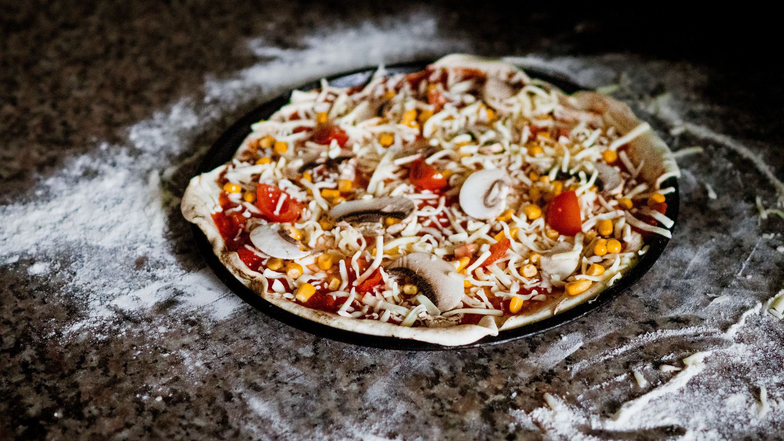 Roher Pizzateig auf einem bemehlten Blech, belegt mit Champignons, Tomaten, Mais und Käse, kurz vor dem Ofen