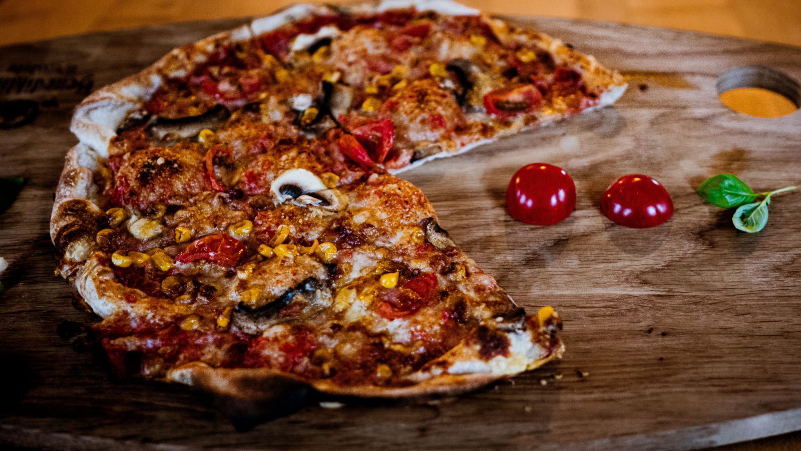 Ofenfrische Pizza mit Champignons, Tomaten und Mais auf einem Holzbrett, ein Stück ist bereits herausgeschnitten